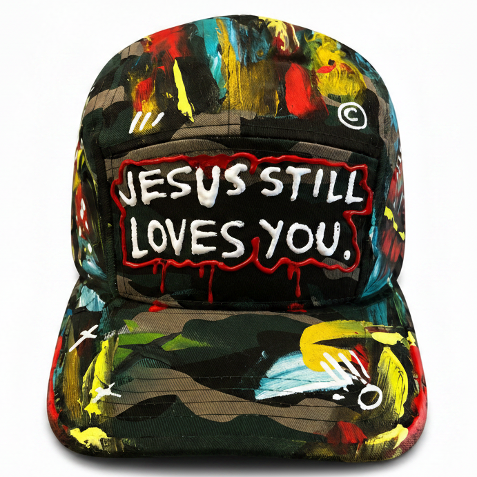 A five-panel baseball cap featuring a colorful abstract paint-splatter pattern in red, yellow, blue, green, and black. The front panel has a central rectangular patch with the text 'JESUS STILL LOVES YOU.