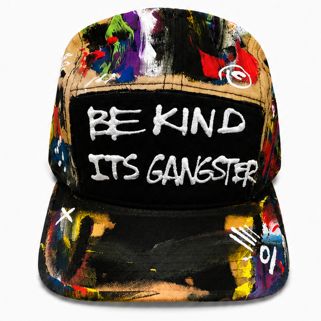A black 5-panel snapback cap with a colorful abstract paint splatter pattern on the panels and brim, featuring a black front patch with the white text 'BE KIND ITS GANGSTER'.