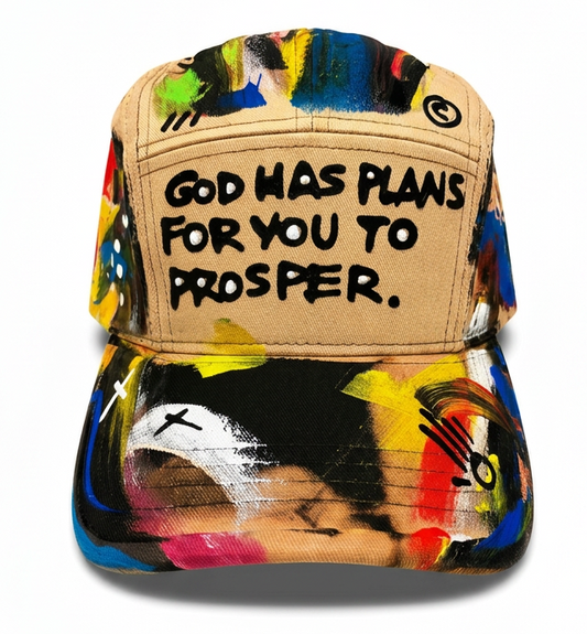 A baseball cap featuring a beige front panel with the text 'GOD HAS PLANS FOR YOU TO PROSPER.' in black lettering, surrounded by a vibrant, multicolored abstract paint-splatter pattern on the brim and crown.
