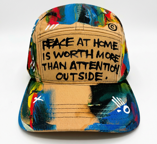 A baseball cap with a beige front panel featuring the text 'PEACE AT HOME IS WORTH MORE THAN ATTENTION OUTSIDE' in black, surrounded by a colorful abstract paint-splatter pattern in blue, green, red, yellow, and black.